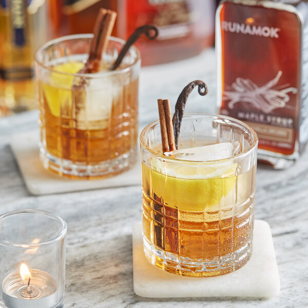 A bottle of Runamok Cinnamon and Vanilla-Infused Maple Syrup on a table with two glasses of brown liquid and cinnamon sticks.