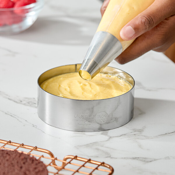 A stainless steel custard ring being filled with batter using a piping bag.