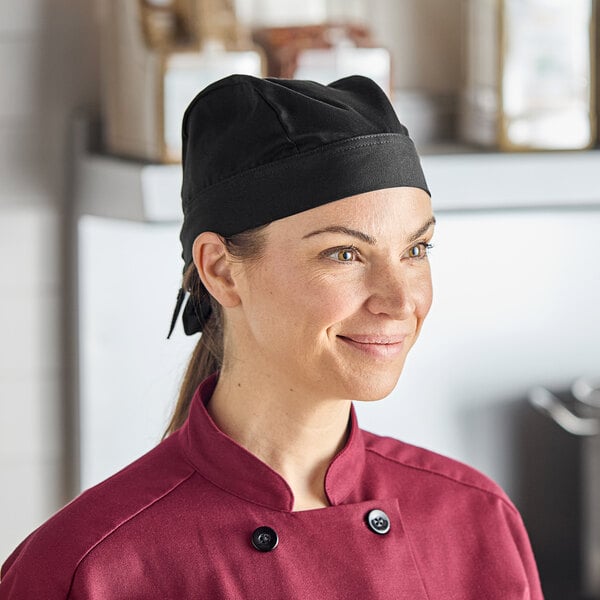 A woman wearing a black Uncommon Chef skull cap with ties smiles in a professional kitchen.