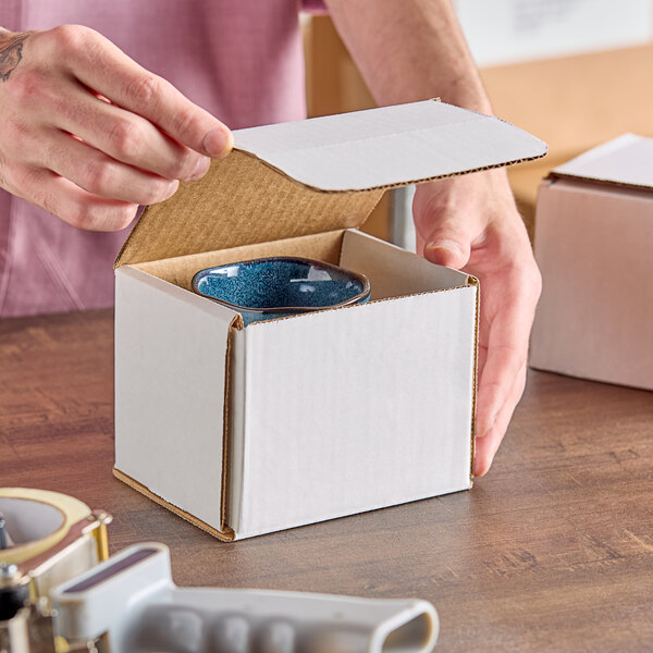 A white corrugated mailer box with a tuck top being packed with a ceramic cup.