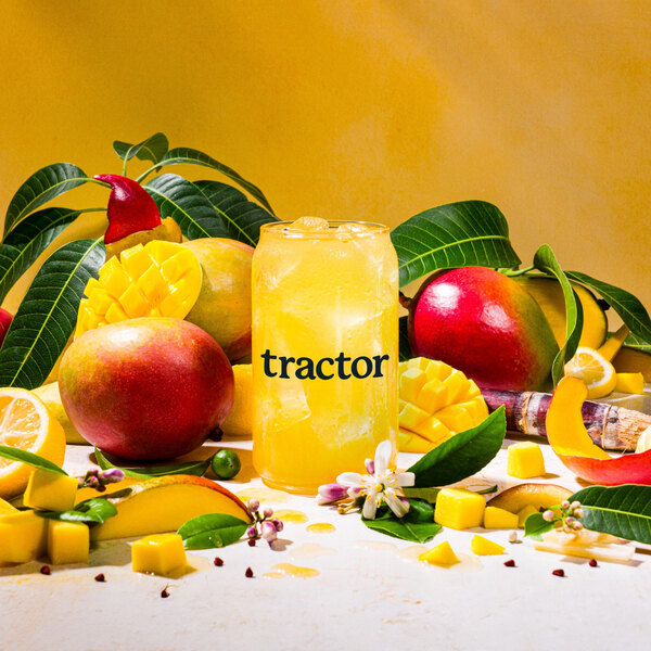 A glass of yellow Tractor Beverage Co. mango liquid with fruit and leaves on the rim.