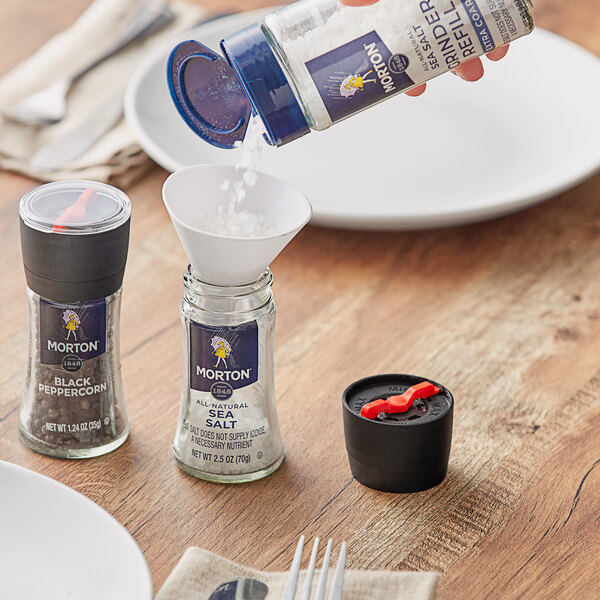 A person refilling a salt shaker with Morton Extra Coarse Sea Salt using a red handled grinder.