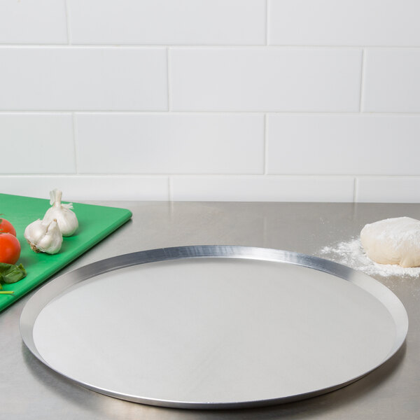 An American Metalcraft heavy weight aluminum pizza pan on a counter with tomatoes, garlic, and basil.