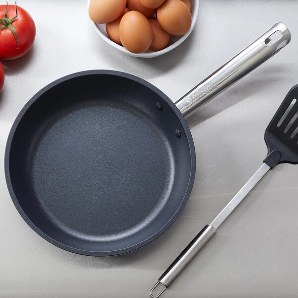 A non-stick fry pan with a stainless steel handle, shown on a countertop with a spatula, eggs, and tomatoes nearby.