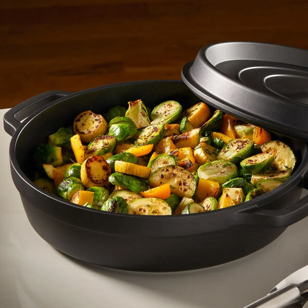 A black non-stick round casserole dish with a cover, filled with roasted vegetables.
