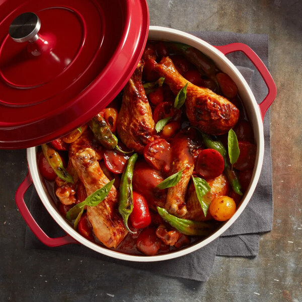 A red non-stick round casserole dish with a cover, filled with cooked chicken drumsticks, vegetables, and herbs.