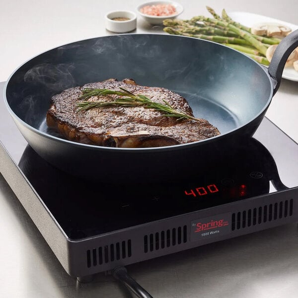 A black carbon steel fry pan with a steak cooking inside, placed on an induction cooktop.