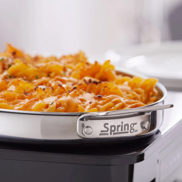 A stainless steel oval saute pan with handles, filled with baked pasta, labeled 'Spring USA Primo! 1.5 Qt. 5-Ply Oval Saute Pan with Stainless Steel Handles.'