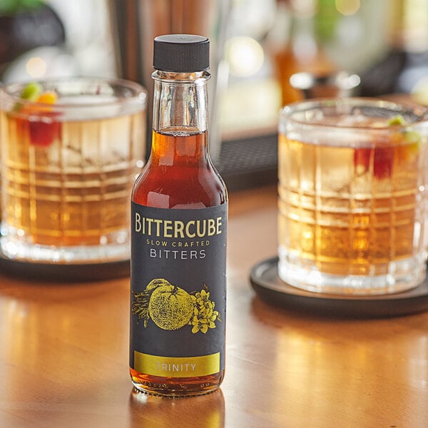 A bottle of Bittercube Trinity Bitters on a table next to two glasses of liquid.