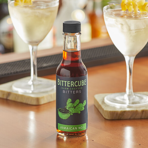 A bottle of Bittercube Jamaican No. 1 Bitters on a table next to two glasses of liquid.