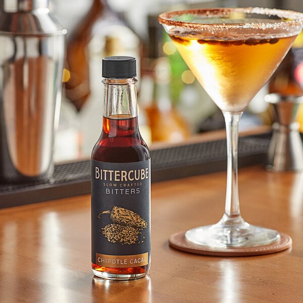 A glass of liquid with a bottle of Bittercube Chipotle Cacao Bitters on a table.