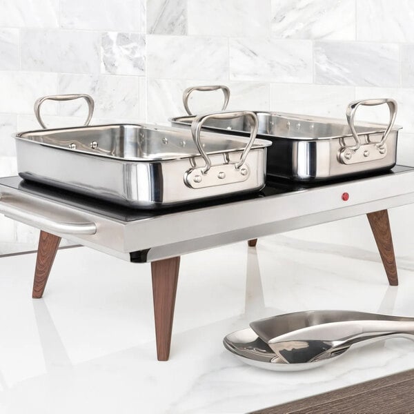 A stainless steel roasting pan with handles, placed on a modern warming tray with wooden legs, accompanied by serving utensils.