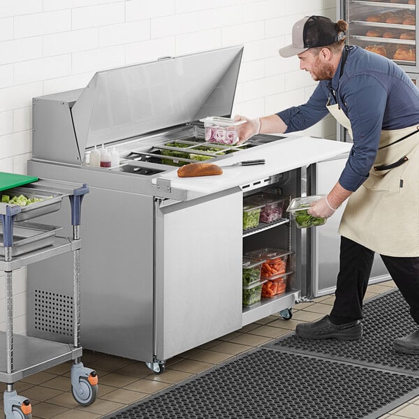 A man in a white apron and black pants using an Avantco stainless steel sandwich prep table to prepare food.