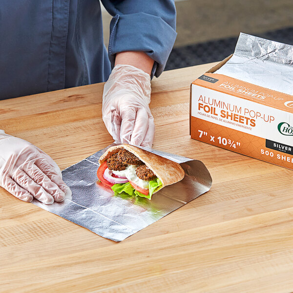 A person in gloves using Choice interfolded foil sheets to wrap a sandwich on a cutting board.