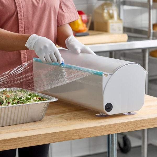 A plastic film and foil dispenser and cutter being used to cover a food tray with plastic wrap.