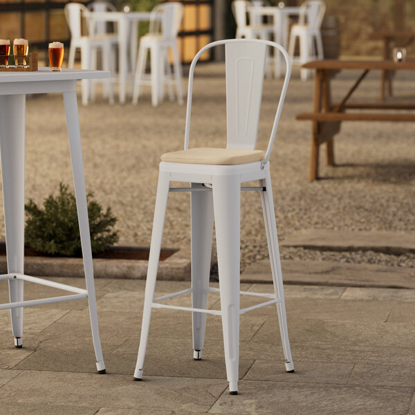 A white Lancaster Table & Seating outdoor cafe barstool with a tan cushion on a table outside.