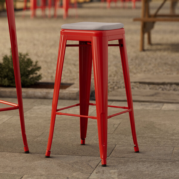 A red Lancaster Table & Seating outdoor barstool with a gray cushion on a stone surface.