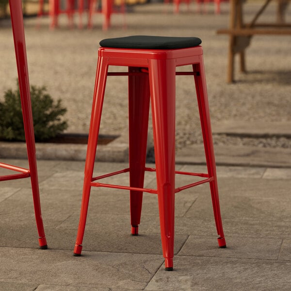 A red Lancaster Table & Seating outdoor bar stool with a black cushion on a stone surface.