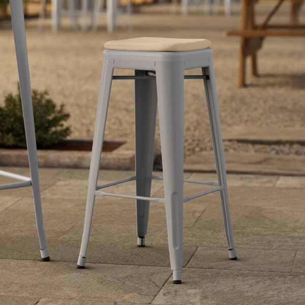 Two Lancaster Table & Seating nickel gray metal outdoor barstools with tan cushions on a stone patio.