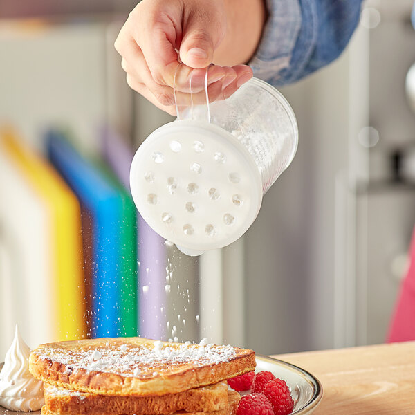 A person pouring coarsely ground cheese from a clear polycarbonate shaker onto a plate of food.