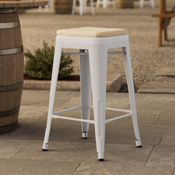 A white Lancaster Table & Seating backless counter height stool with a tan cushion on a stone surface.