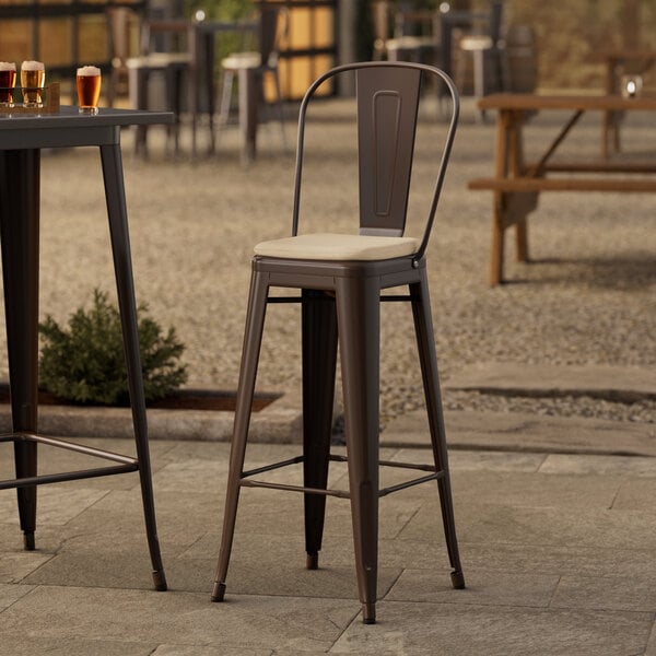 A Lancaster Table & Seating copper outdoor barstool with a tan cushion on an outdoor patio table.