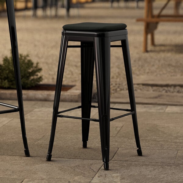 A black Lancaster Table & Seating outdoor backless barstool with a black cushion on a stone surface.