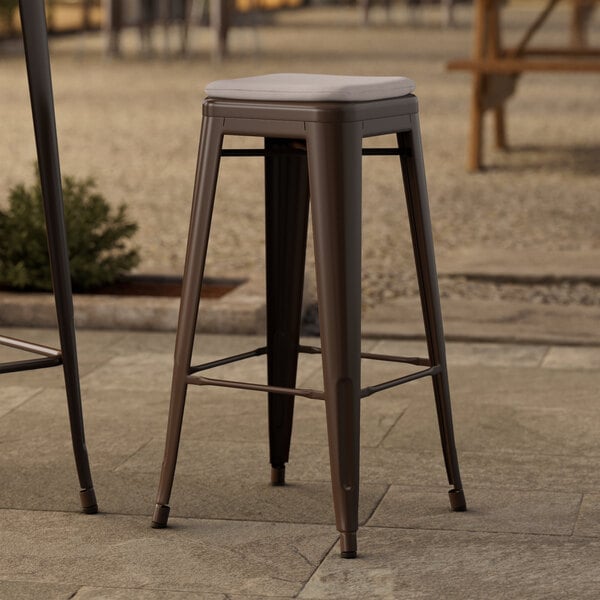 Two Lancaster Table & Seating copper outdoor barstools with gray cushions on a stone patio.