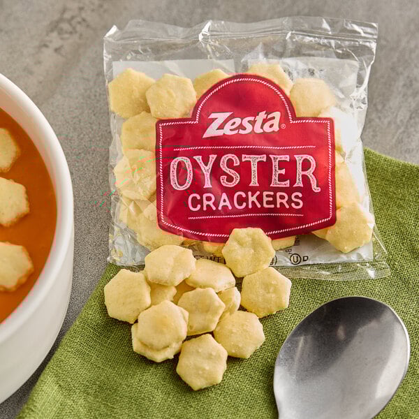 A small bag of Zesta oyster crackers with some crackers spilled out next to a spoon and a bowl of soup.