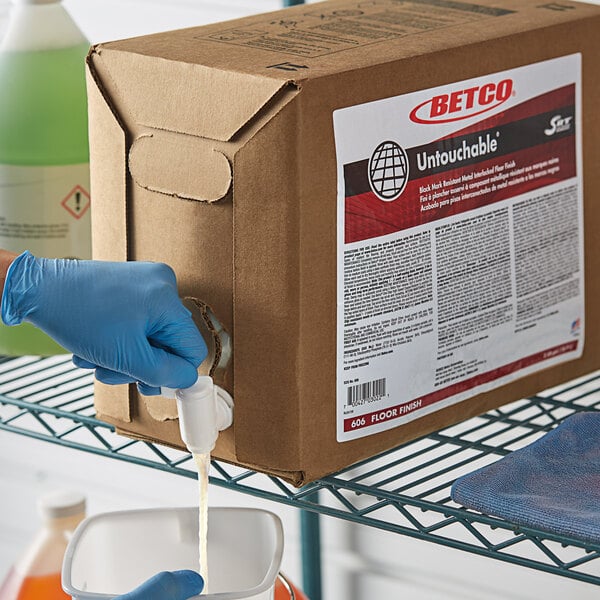 A person wearing blue gloves pouring Betco floor finish from a bottle into a box.