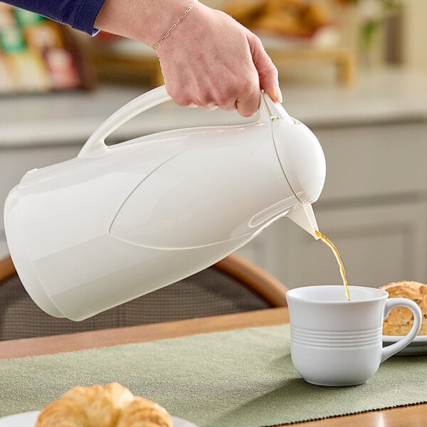 A person pouring coffee from a white Zojirushi glass carafe into a white mug.