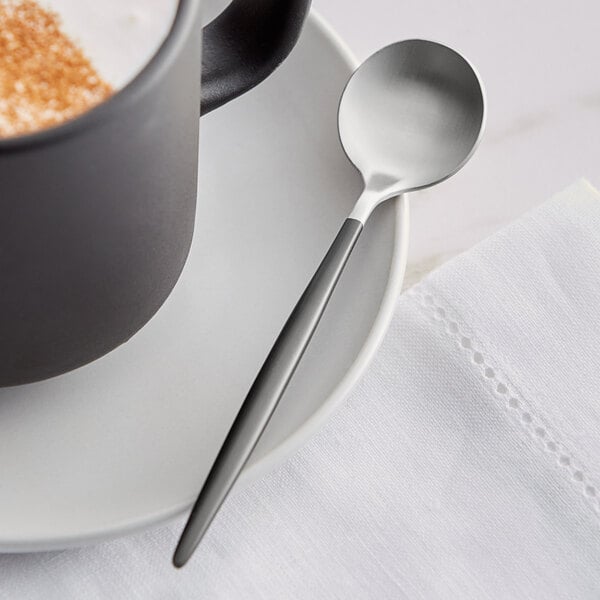 An Acopa Odin stainless steel demitasse spoon on a saucer with a cup of coffee.