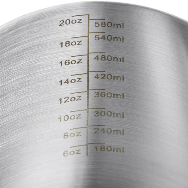 A stainless steel Estella Caffe frothing pitcher with measurements on a counter.