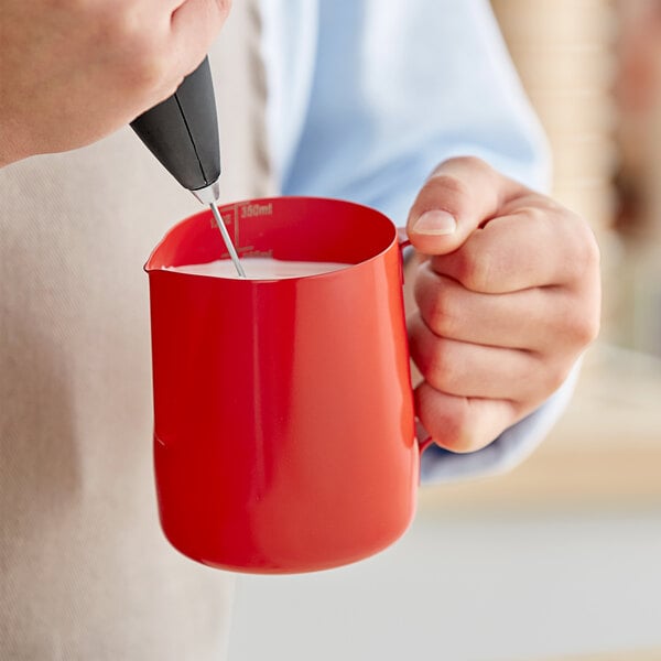 A person using a red Acopa frothing pitcher to pour milk into a mug.