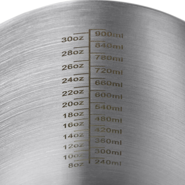 A stainless steel Estella Caffe frothing pitcher with measurements on a table.