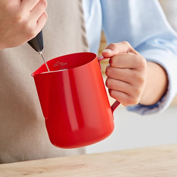 A person using an Acopa red frothing pitcher to pour milk into a red cup.