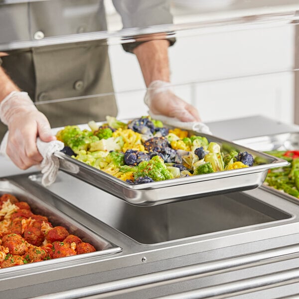 A person in a white uniform holding a Vollrath stainless steel steam table pan filled with food.