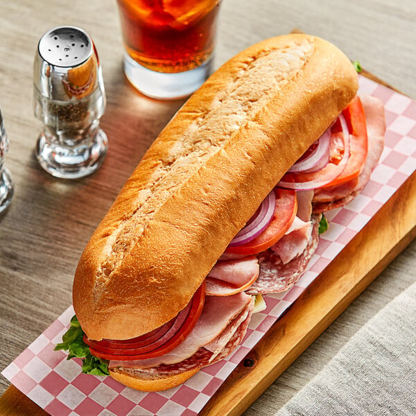 A sandwich with meat, tomatoes, and onions on a Turano Soft Sub Roll on a cutting board.