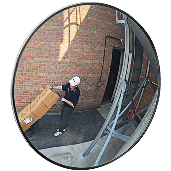 A man holding a cart with a stack of boxes in front of a Vestil outdoor convex mirror.