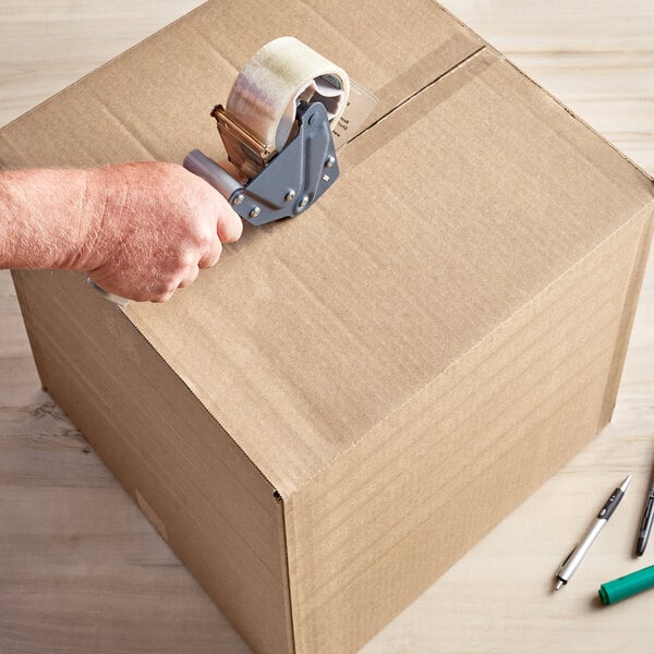 A hand using a tape dispenser to seal a Lavex cardboard shipping box.