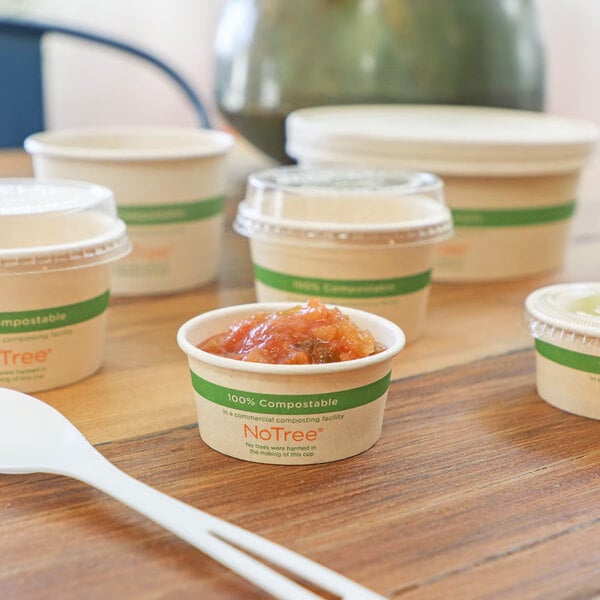 A small compostable portion cup filled with sauce, surrounded by other similar cups and a white plastic utensil on a wooden table.