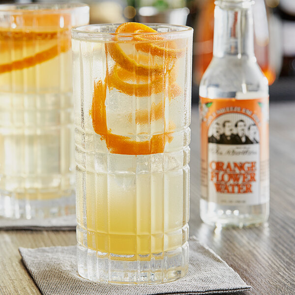 A glass of liquid with orange slices and a bottle of Fee Brothers Orange Flower Water on a table in a cocktail bar.