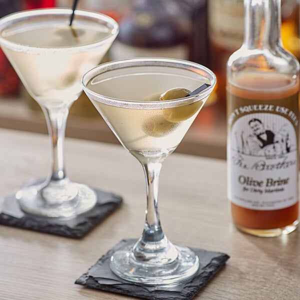 A pair of martini glasses with Fee Brothers Olive Juice and olives on a table.