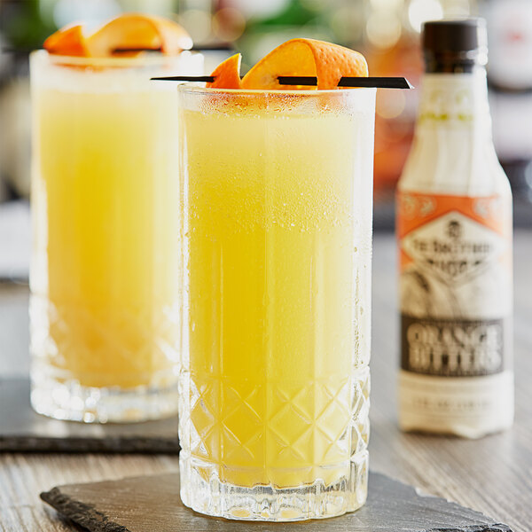 A bottle of Fee Brothers Gin Barrel Aged Orange Bitters on a table with two glasses of orange juice and a bottle of liquor.