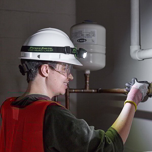 A woman wearing a hard hat and safety glasses using a PowerSmith LED headlamp to work on a pipe.