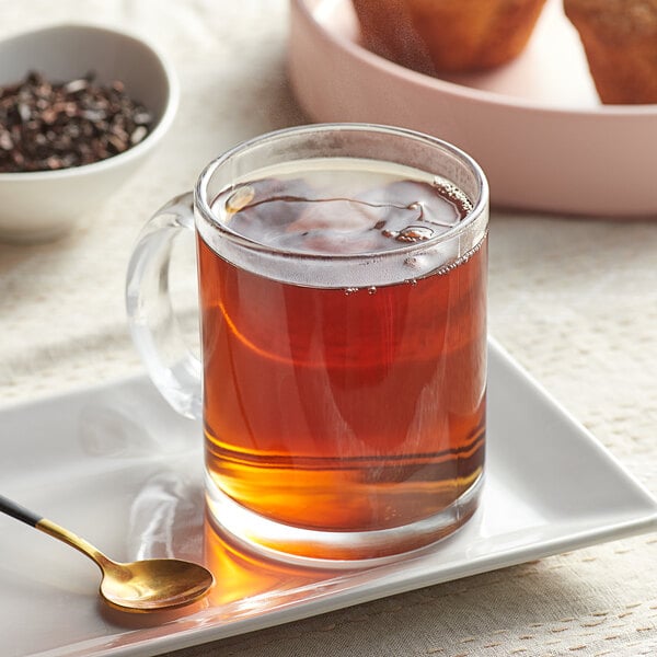 A glass mug of Davidson's Organic Quilan China Oolong tea on a white plate with a bowl of muffins and a spoon.