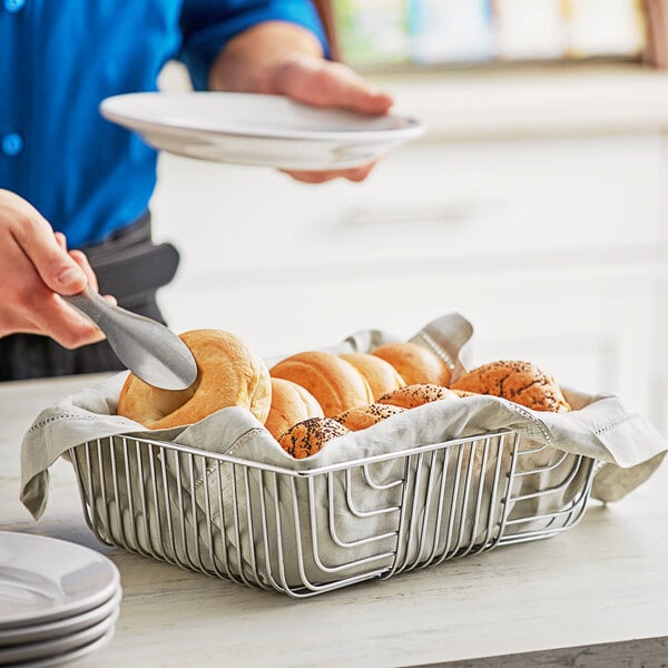 A person putting a spoon into a rectangular wire basket filled with bagels.