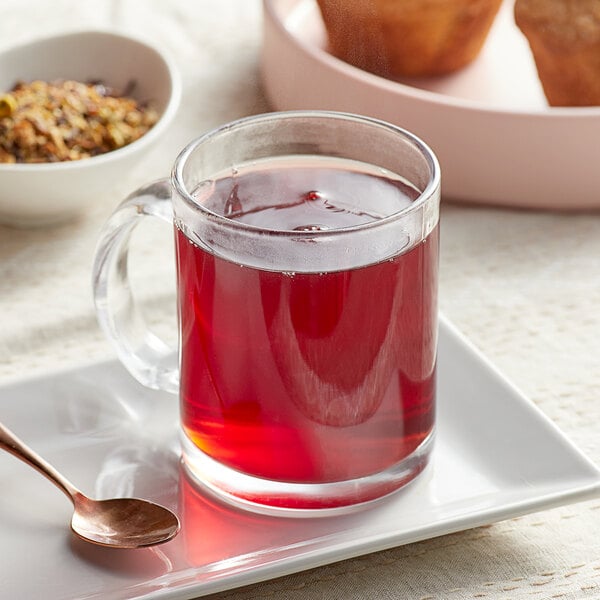A glass mug of Davidson's Organic Garden Harvest Herbal tea on a white plate with a spoon and muffins.