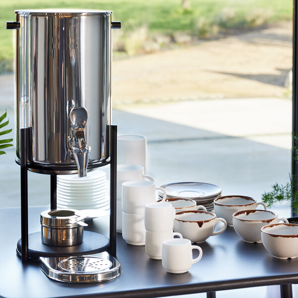 A white table with an Abert Cosmo stainless steel coffee urn and white mugs filled with coffee.