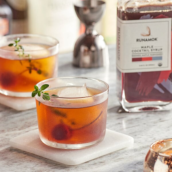 Two glasses of Runamok Smoked Maple Old Fashioned cocktails with cherries and herbs on a counter.